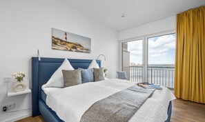 A modern bedroom with a large bed, a view of the sea, and a painting of a lighthouse.
