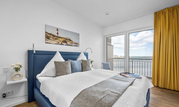 A modern bedroom with a large bed, a view of the sea, and a painting of a lighthouse.