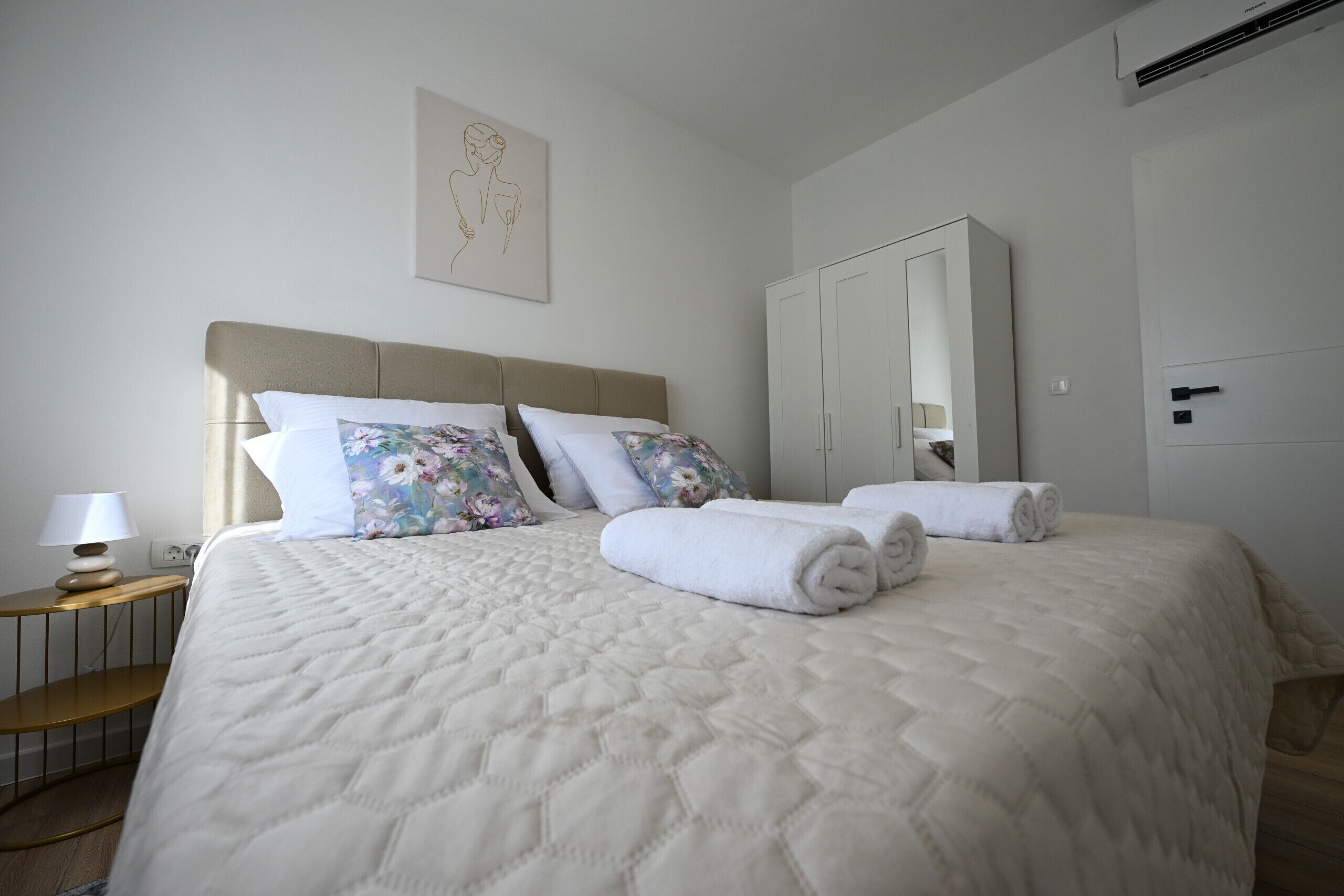 Ein ordentlich bezogenes Bett mit einer gesteppten weißen Daunendecke, zwei blumigen Kissen und gerollten weißen Handtüchern.