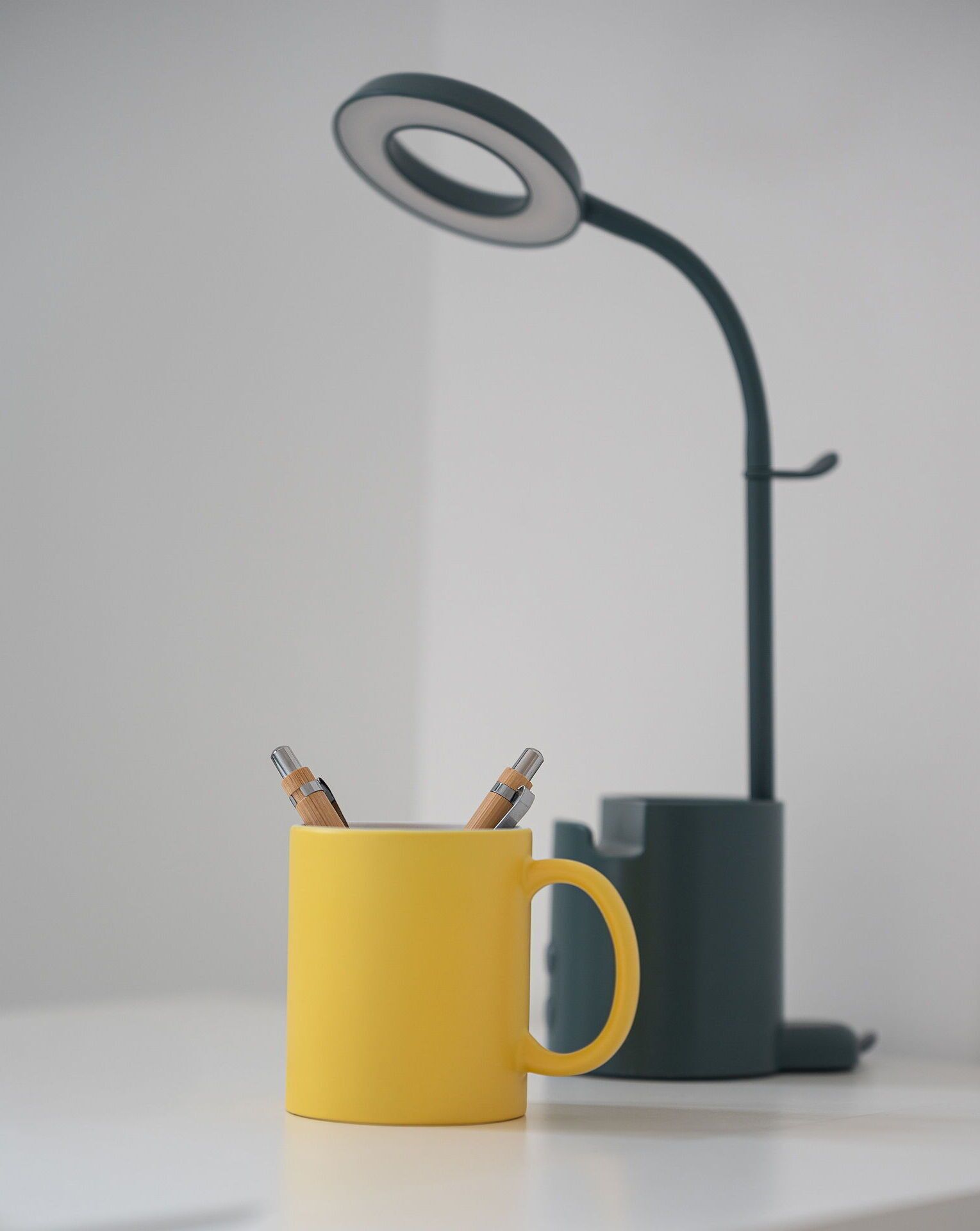 Une tasse jaune avec des stylos posés sur une surface blanche, une lampe de bureau noire et un objet gris à l’arrière-plan.