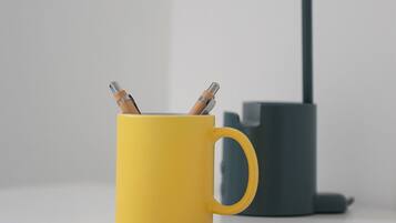 Une tasse jaune avec des stylos posés sur une surface blanche, une lampe de bureau noire et un objet gris à l’arrière-plan.