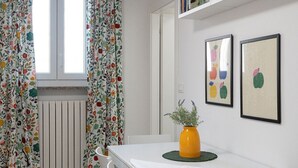 A dining area with a white table, chairs, and a vase with flowers. There are two framed pictures on the wall and a window with curtains. - Lovely apartment for 4 guests with A/C, WIFI and TV (Pavia)