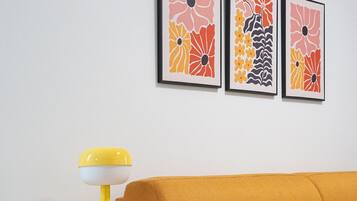 Een kamer met een mosterdkleurige bank, een kleine tafel met een gele lamp, en drie lijstwerken met abstracte kunst aan de muur.