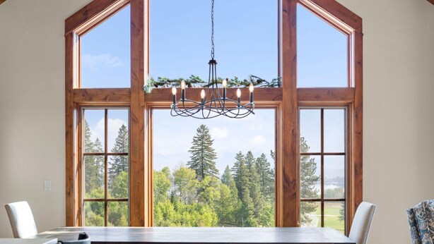 Un espace repas avec une grande table et des chaises en bois, un lustre et une vue sur une forêt à travers de grandes fenêtres.