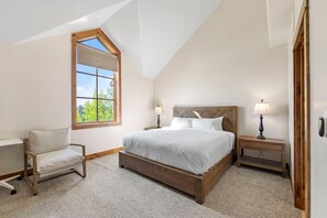 A bedroom with a sloped ceiling, a large bed with a wooden headboard, two bedside tables with lamps, a chair, and a window with a view of greenery.