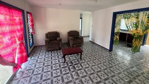 Een kamer met twee bruine fauteuils, een houten salontafel, vloertegels met een patroon, en een raam met roze gordijnen.