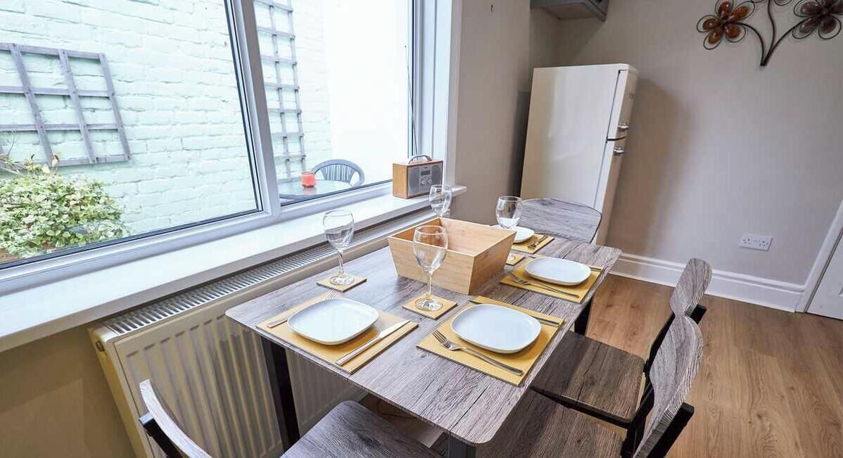 Un coin repas avec une table dressée pour quatre personnes, un radiateur, une fenêtre et un appareil électroménager blanc en arrière-plan.