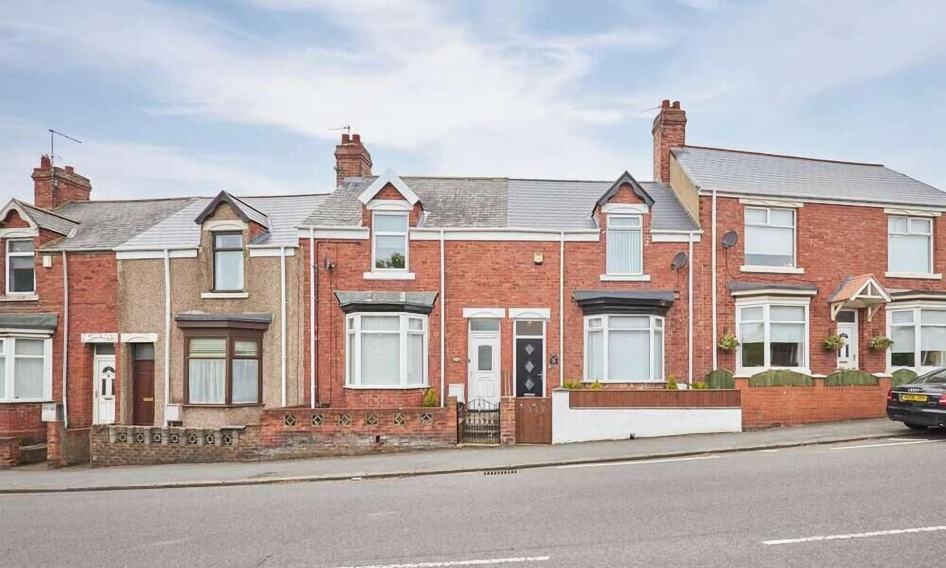 A row of red brick terraced houses with pitched roofs, front doors, and small front gardens.