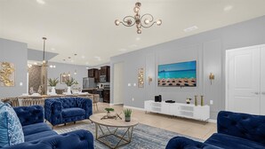 A modern living room with a large TV, blue sofas, a dining area, and a kitchen. - Luxury 7BR Villa w/Pool & Resort Access, Sleeps 16 – 10 Min to Disney (Davenport)