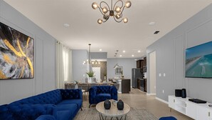 A modern living room with a sectional sofa, a round coffee table, and a dining area with a chandelier. - Luxury 7BR Villa w/Pool & Resort Access, Sleeps 16 – 10 Min to Disney (Davenport)
