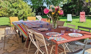 An outdoor dining area with a long table set for multiple people, surrounded by chairs, and a vase with flowers in the center.