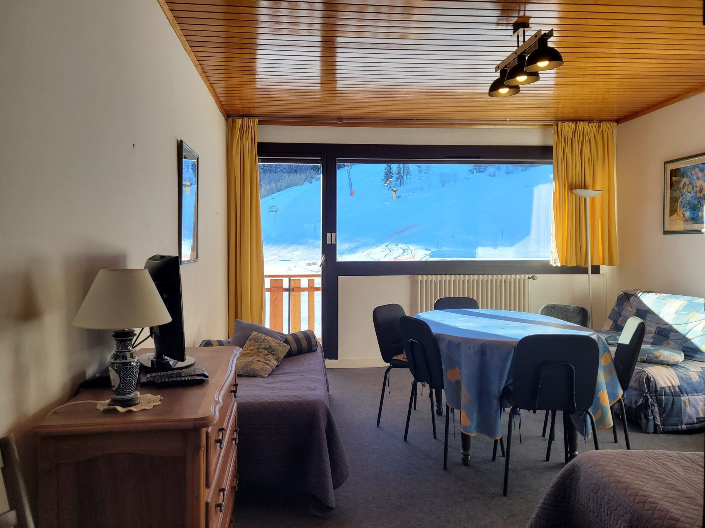 Une pièce chaleureuse avec un plafond en bois, une table à manger avec des chaises, un canapé, une lampe et une vue sur un paysage enneigé par une grande fenêtre.