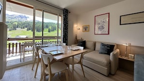 A modern living room with a dining area, large windows offering a mountain view, a sofa, and a framed artwork on the wall.