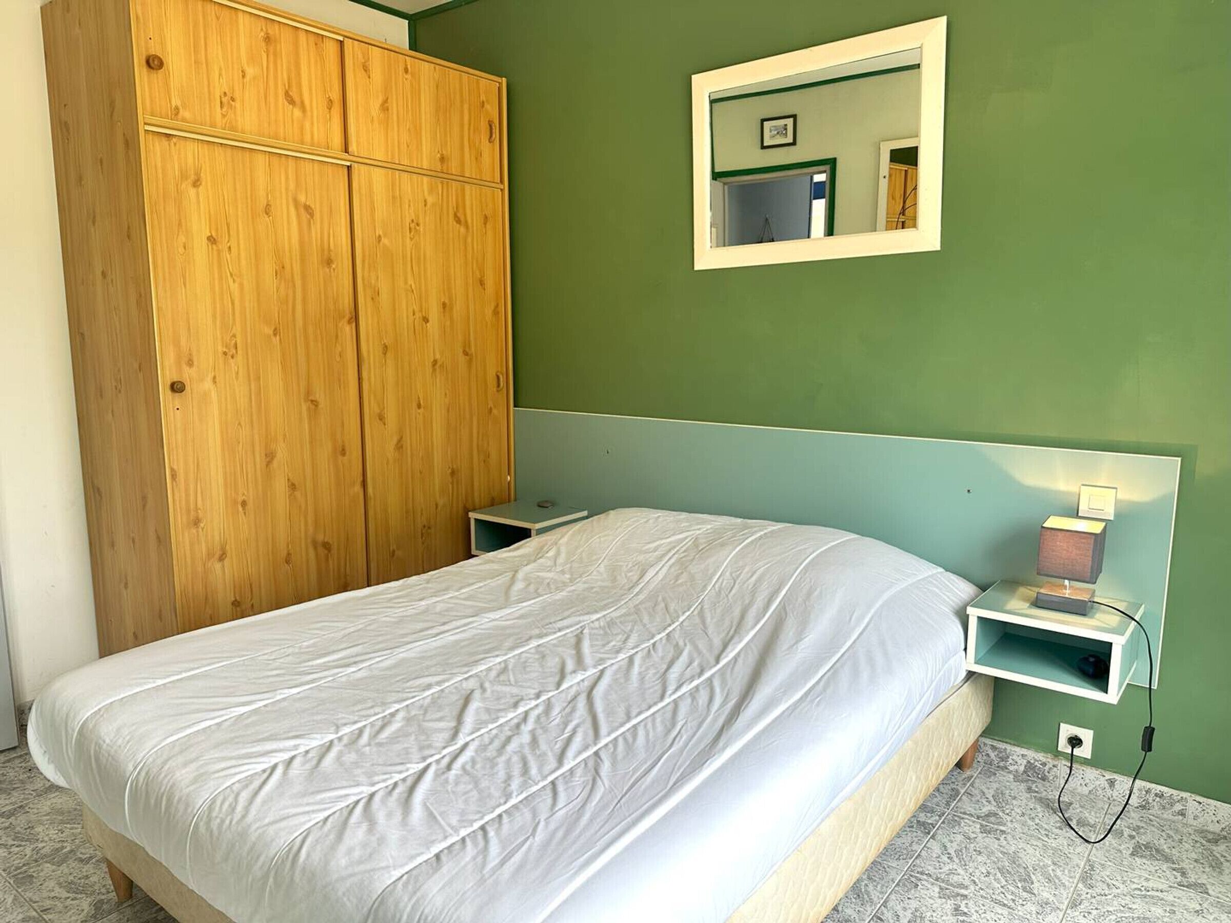 A bedroom with a bed, a wooden wardrobe, a bedside table, and a nightstand.
