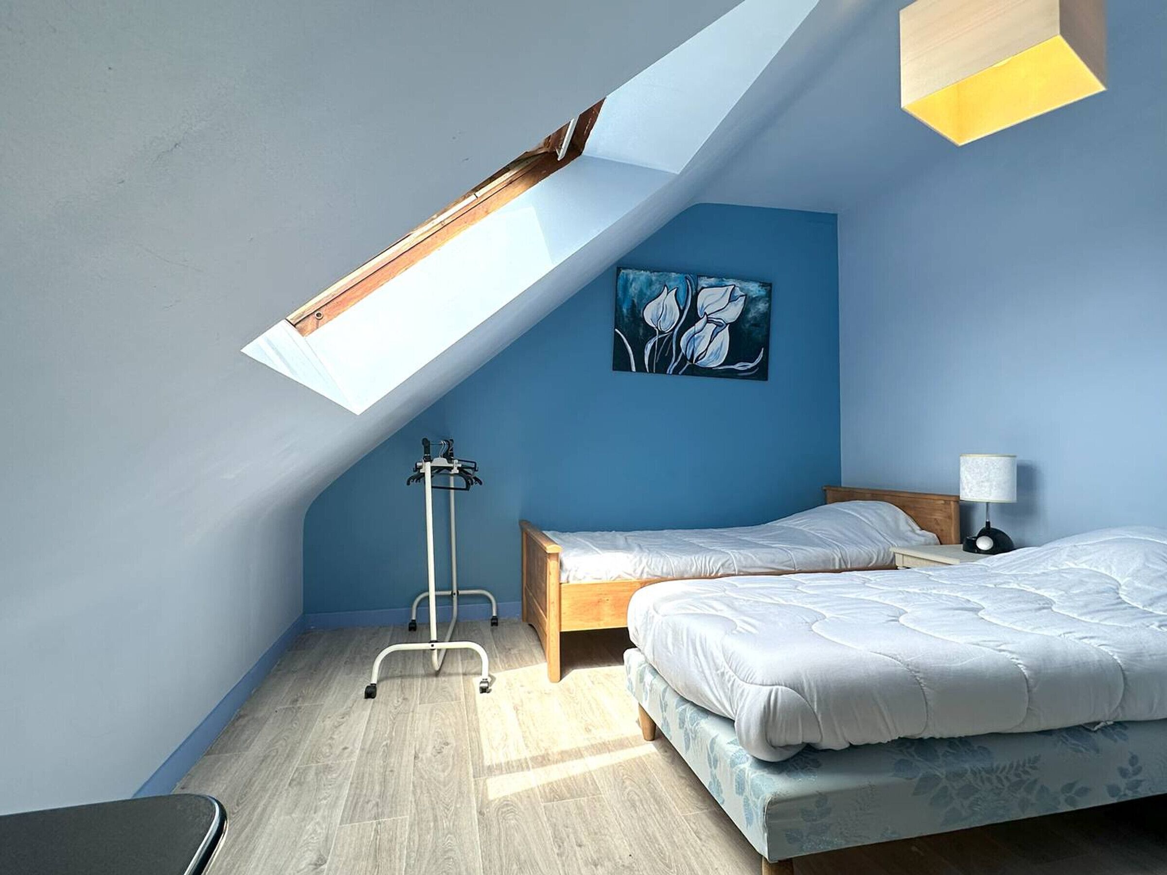 A bedroom with a skylight, two beds, a nightstand, a lamp, and a painting on the wall.