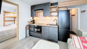 A compact kitchen with a built-in oven, a sink, and a refrigerator. There is a bunk bed with a wooden frame and a small round ottoman. - Ski-in/Ski-out Apartment with Twin Balconies and Free WIFI (Le Corbier)