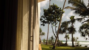 A room with a large window overlooking a grassy area with palm trees and the ocean. There are two wooden chairs and a bench near the window. - Neem Boutique Hotel (Kamburugamuwa)