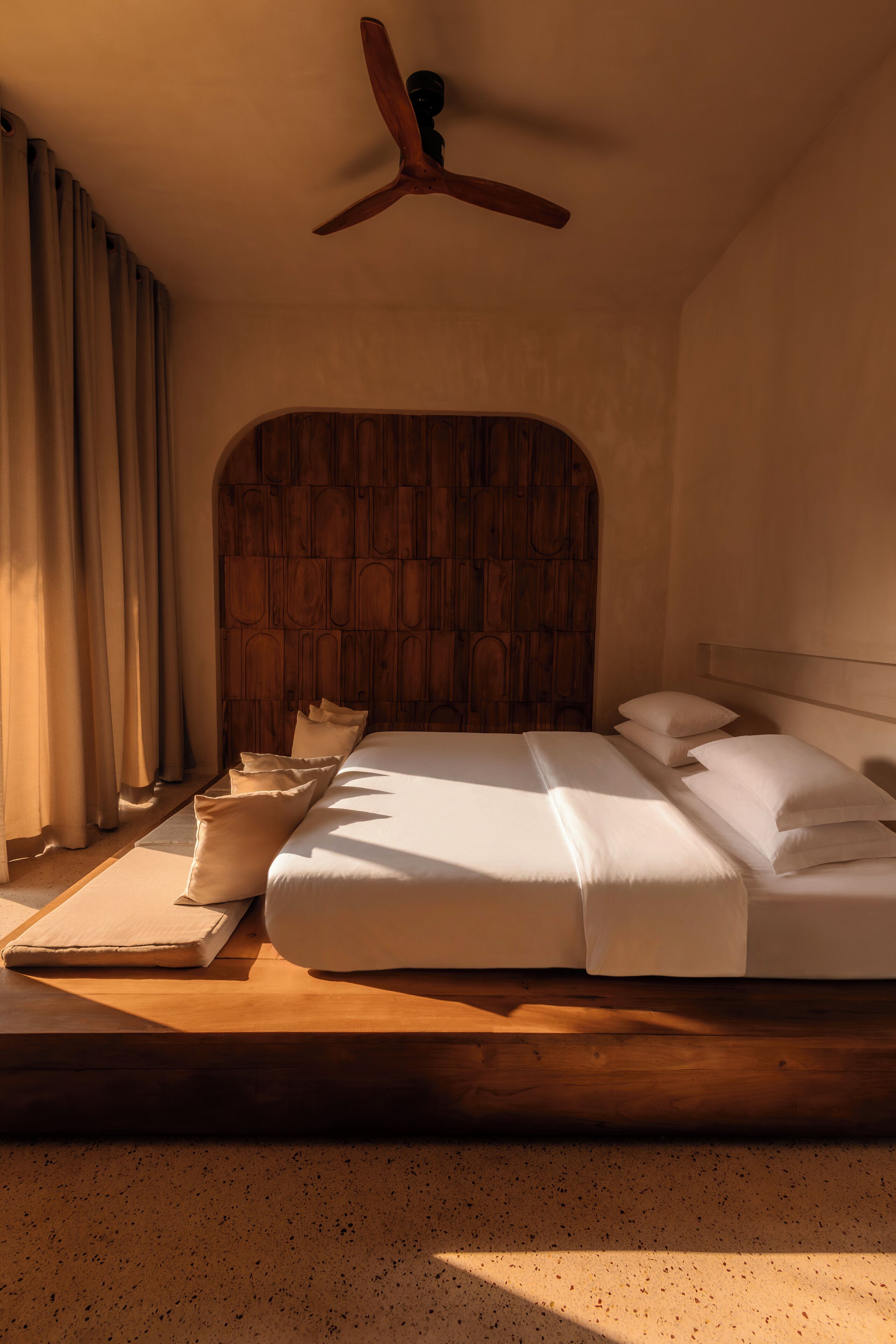 Une chambre à coucher avec une tête de lit en bois, un lit recouvert de draps blancs, un plancher en bois et un ventilateur de plafond.