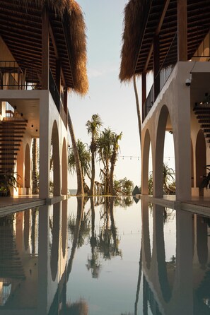 Una piscina con chiari riflessi di palme e un edificio con strutture ad arco e un tetto di paglia.