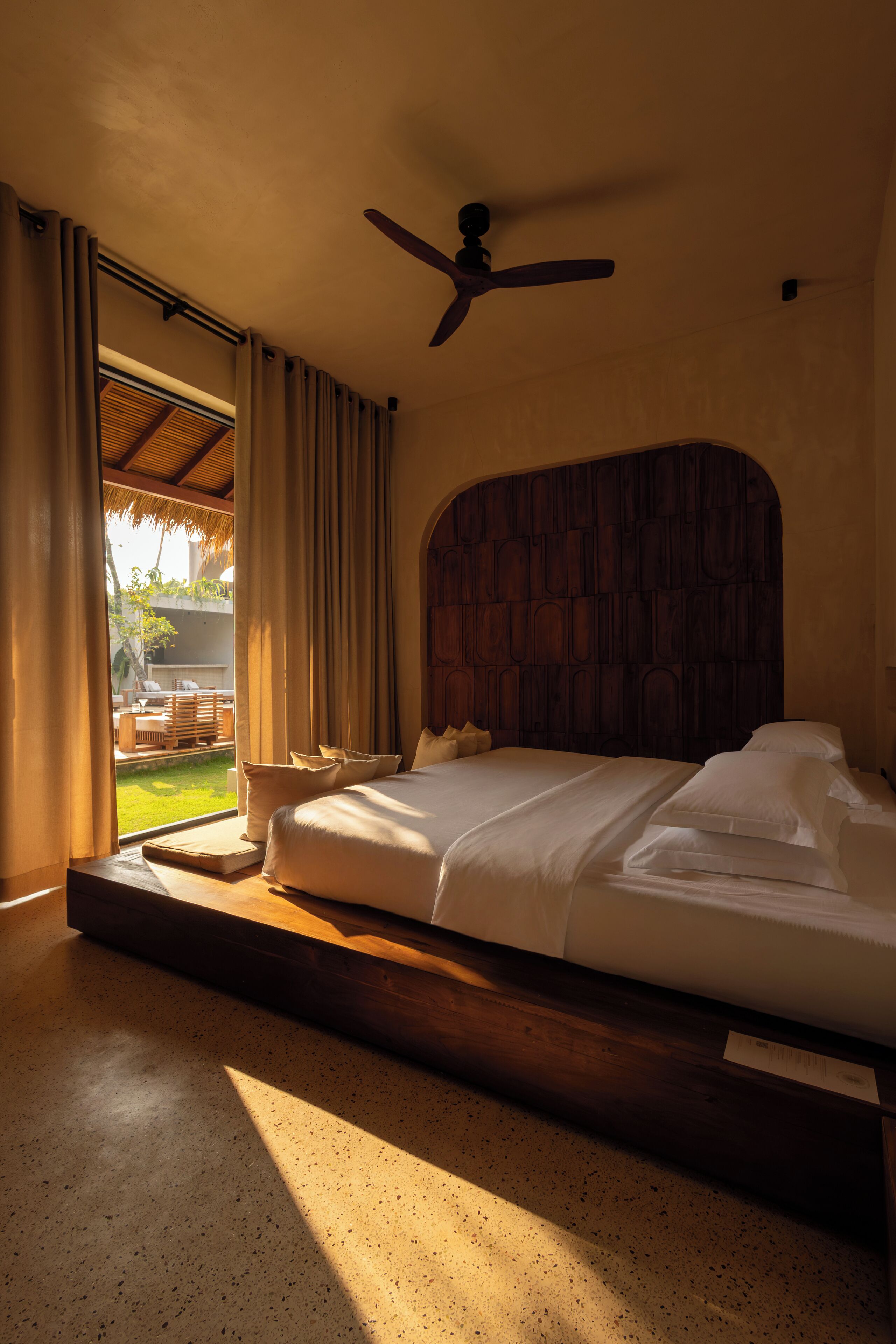 A bedroom with a wooden headboard, a bed with white linens, a ceiling fan, and a view of a patio with outdoor furniture.