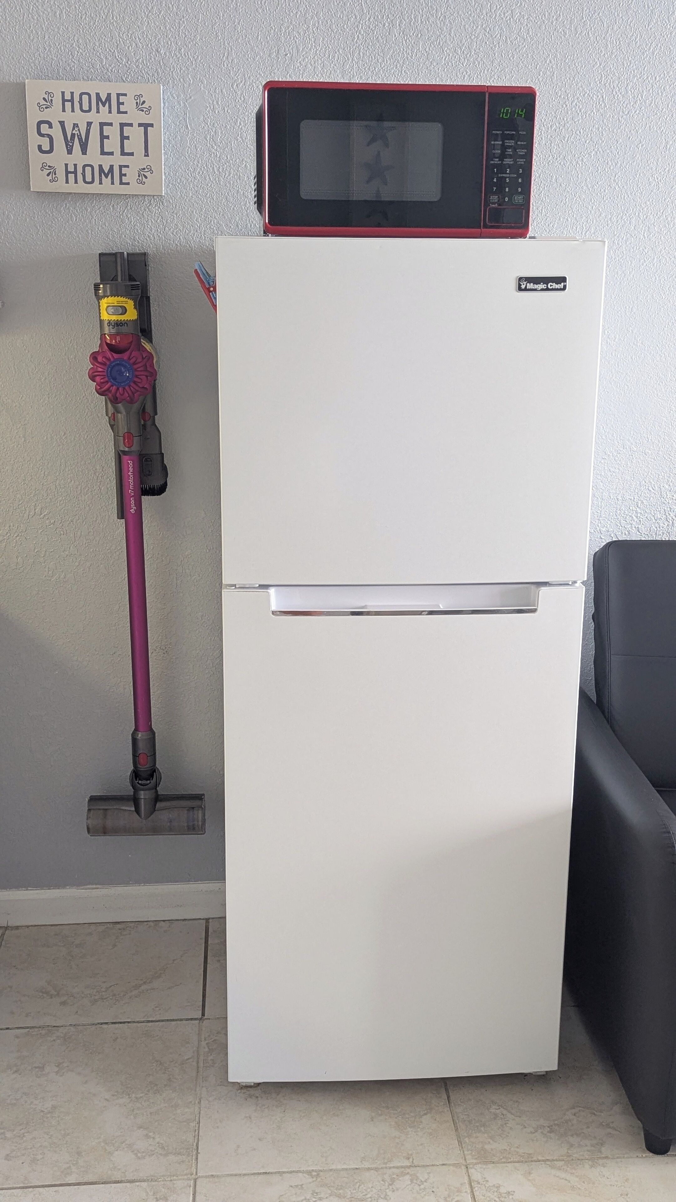 Ein weißer Kühlschrank mit einer Mikrowelle darüber, ein Dyson-Staubsauger und ein Schild an der Wand.