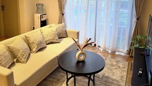 A modern living room with a beige sofa, patterned cushions, a round coffee table with decorative items, and a large window with sheer curtains.