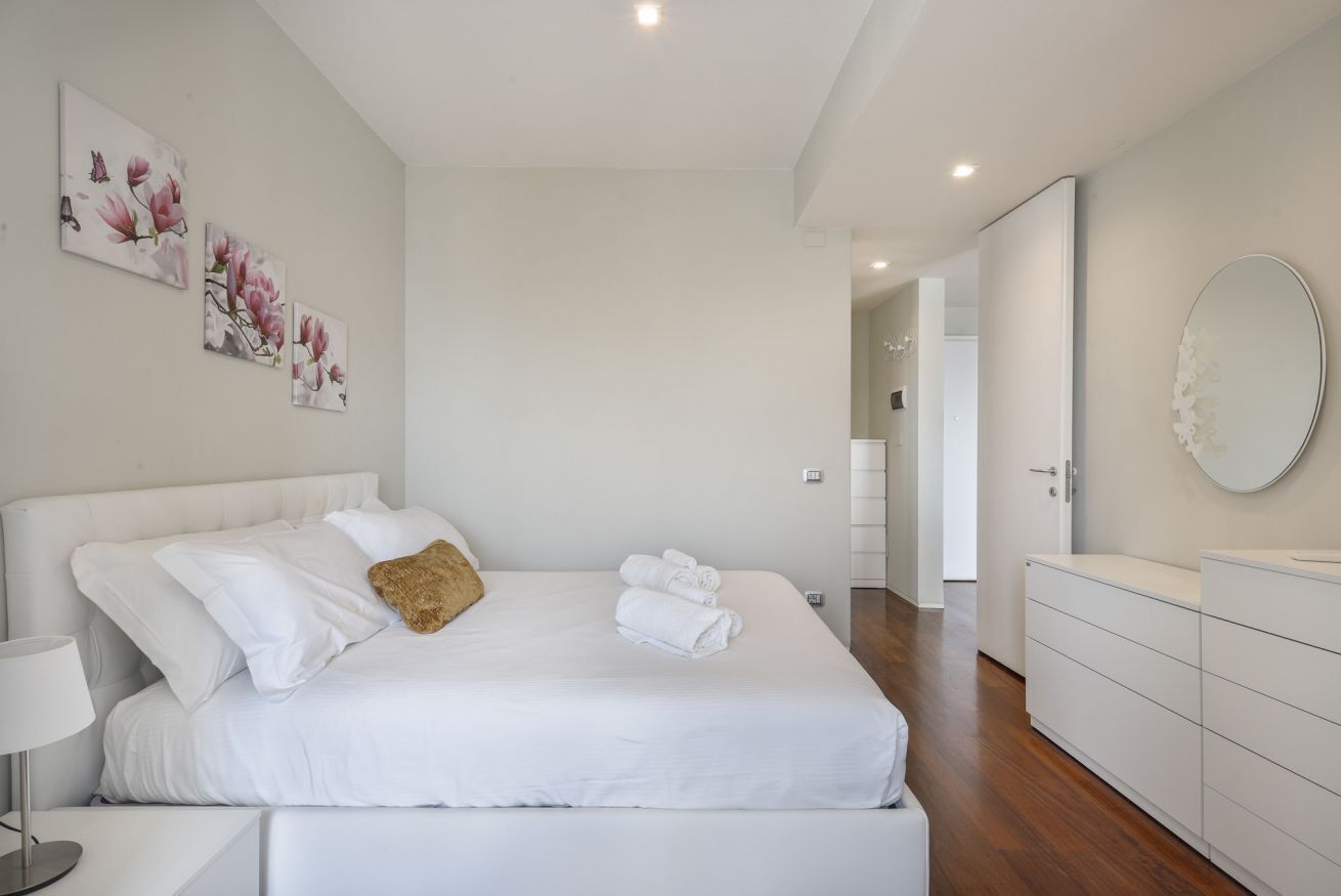 A modern bedroom with a large bed, white linens, a bedside table with a lamp, and a dresser with a mirror.