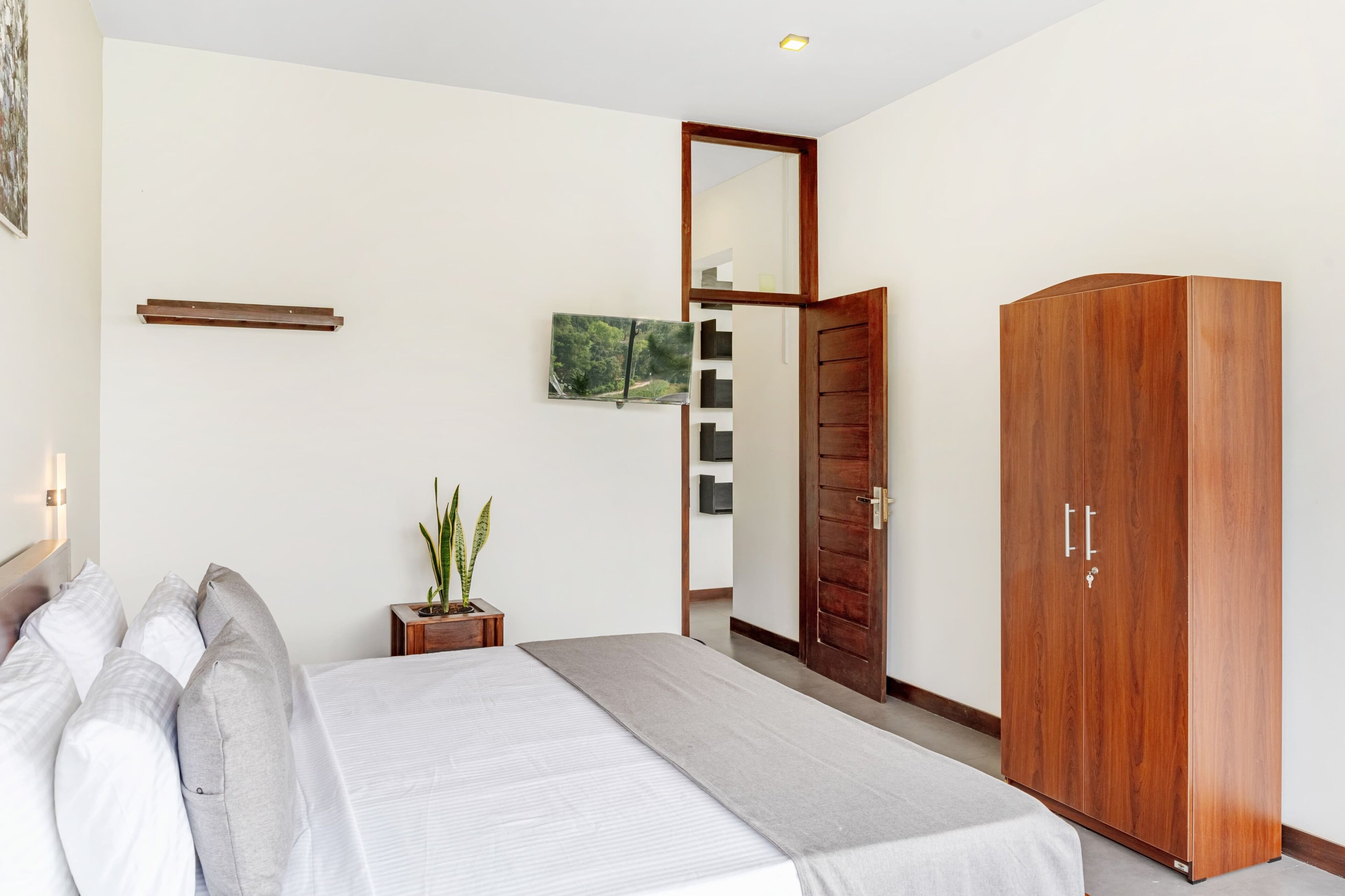 A bedroom with a bed, a wardrobe, a mirror, a small table with a plant, and a wall-mounted shelf.