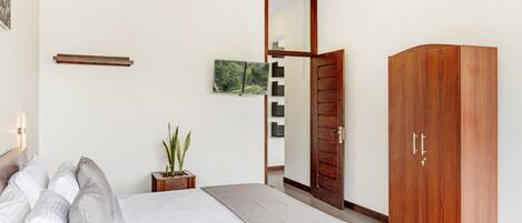 A bedroom with a bed, a wardrobe, a mirror, a small table with a plant, and a wall-mounted shelf.
