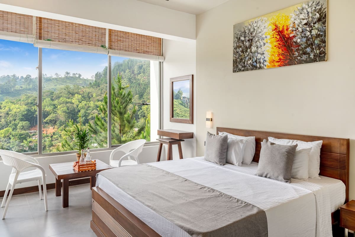 Quarto de hotel com uma janela grande, uma cama, uma escrivaninha, uma cadeira e um quadro na parede.