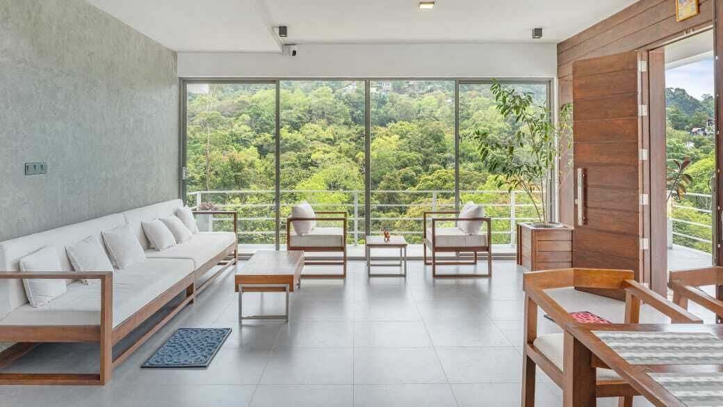 A modern living room with a sofa, chairs, and a table, large windows with a view of greenery, and a wooden door.