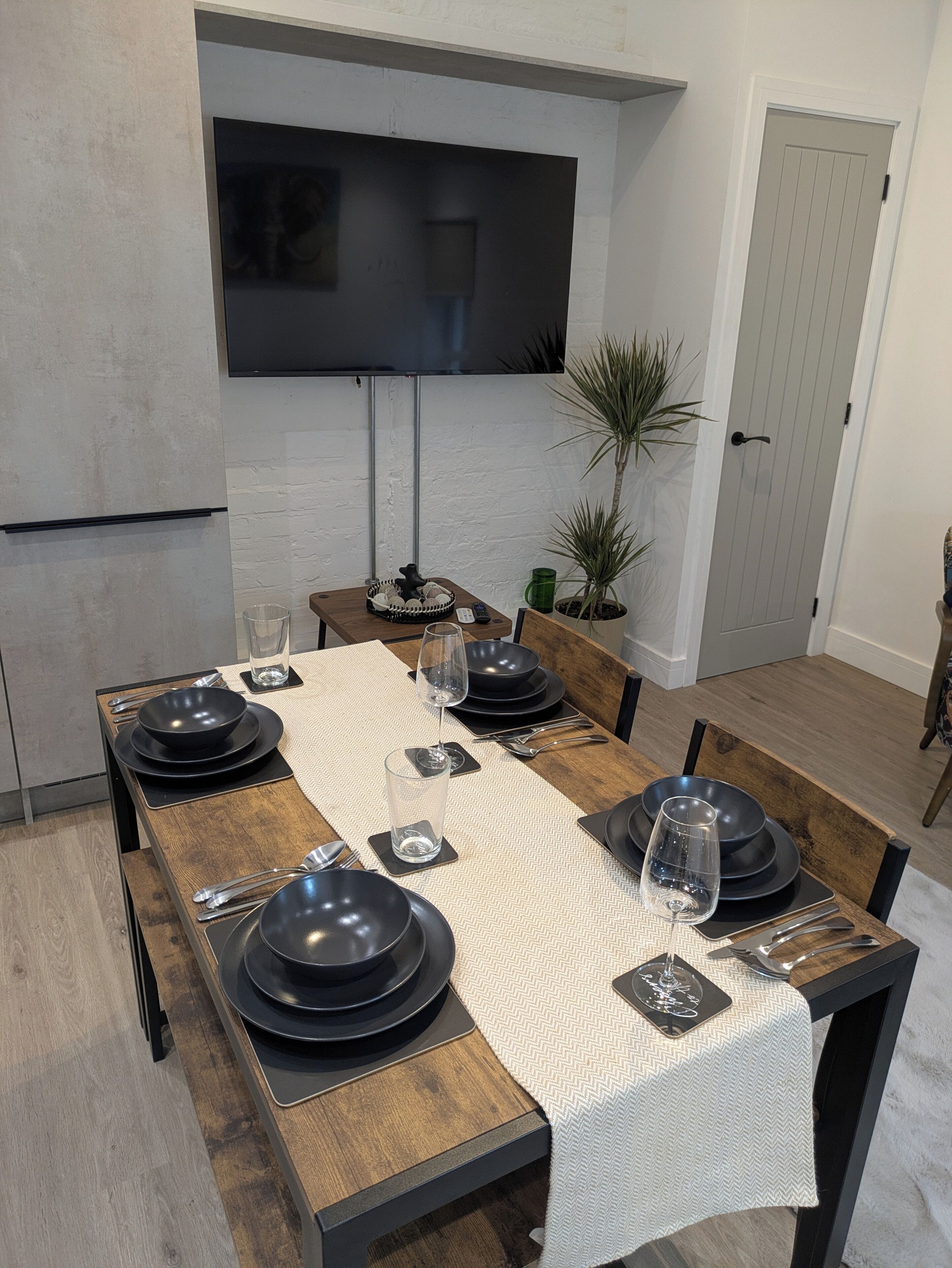 A dining area with a long wooden table set for six, a flat-screen TV mounted on the wall, and a potted plant beside the door.