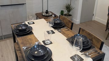 A dining area with a long wooden table set for six, a flat-screen TV mounted on the wall, and a potted plant beside the door.