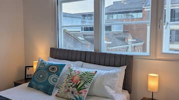 A hotel room with a bed, two bedside lamps, a nightstand, and a window with a view of buildings.