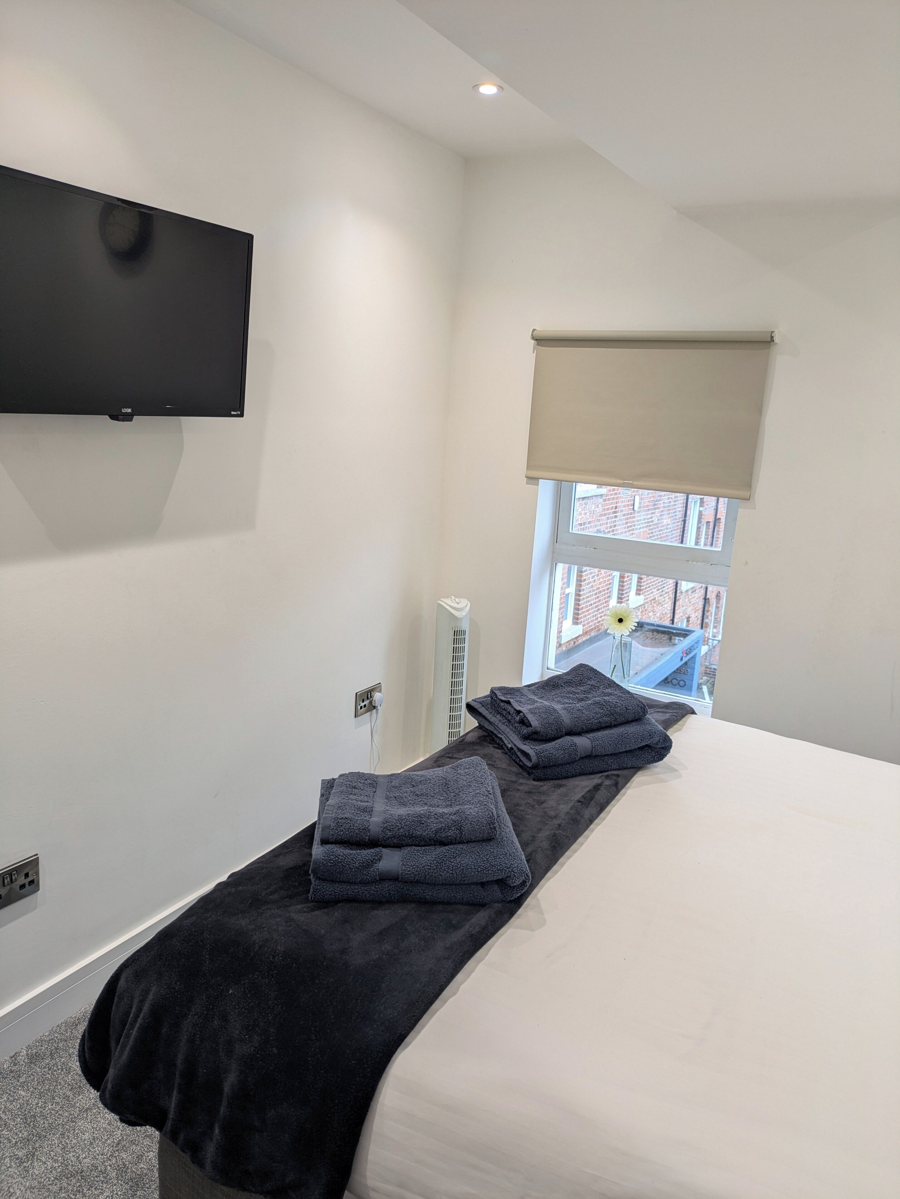 Ein Hotelzimmer mit einem Bett, einem flachbildfernseher an der Wand, einem Fenster mit einer beigen Jalousie und einem kleinen Tisch mit Handtüchern.