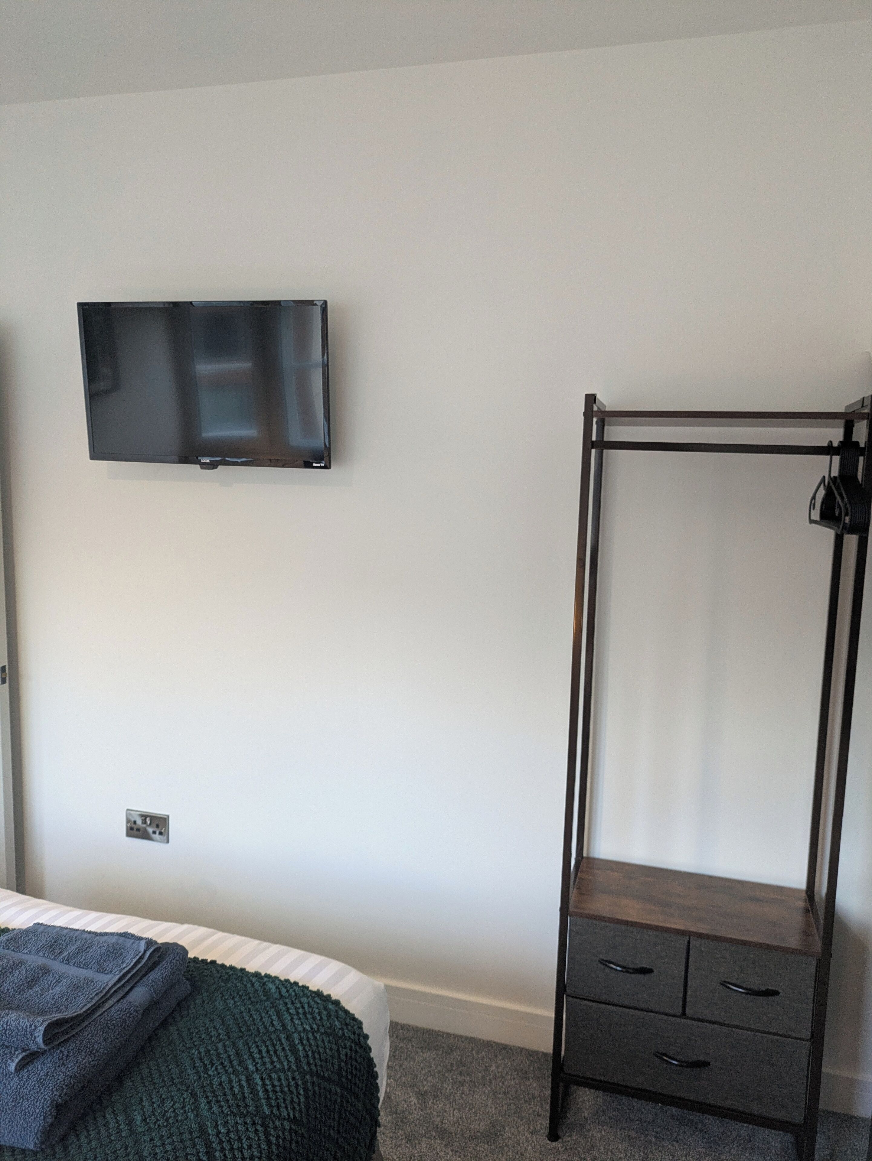 Una habitación con una cama, un televisor montado en la pared y un armario de madera con cajones.