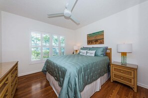 Un dormitorio con cama, lámparas de noche, ventilador de techo, ventana con persianas y un cuadro en la pared.