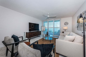 A modern living room with a flat-screen TV, a ceiling fan, and a large window with blinds.