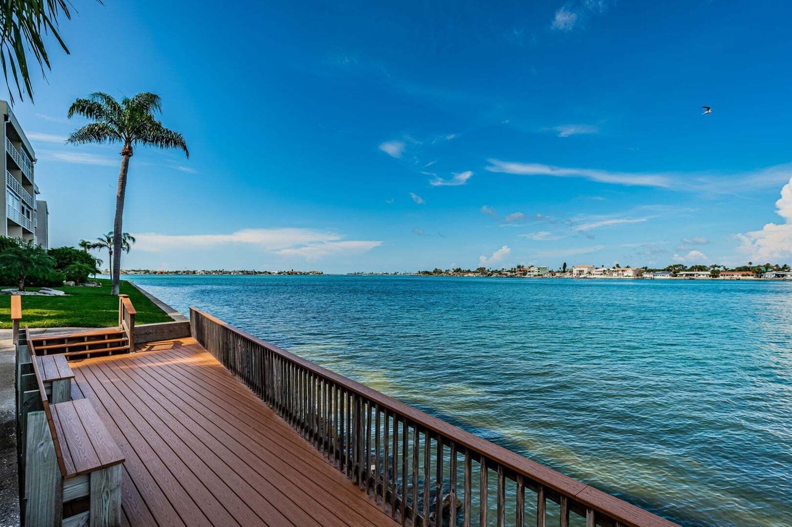 A wooden deck with a railing overlooking a body of water, palm trees, and a distant shoreline with buildings.