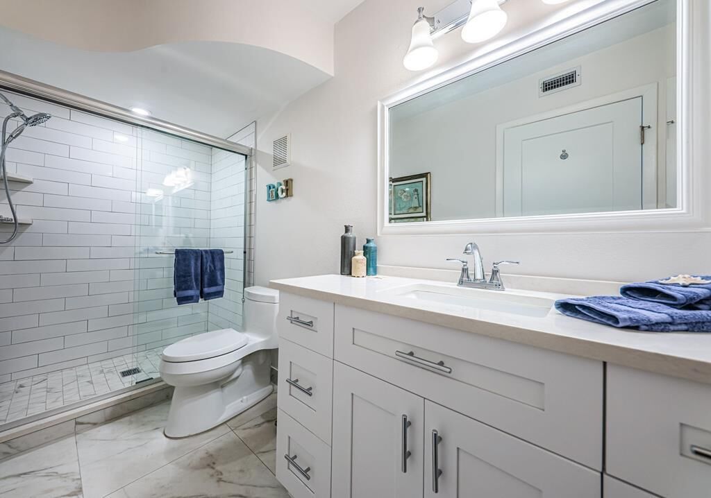 A modern bathroom with a glass shower enclosure, a white toilet, a large mirror, and a white vanity with drawers.
