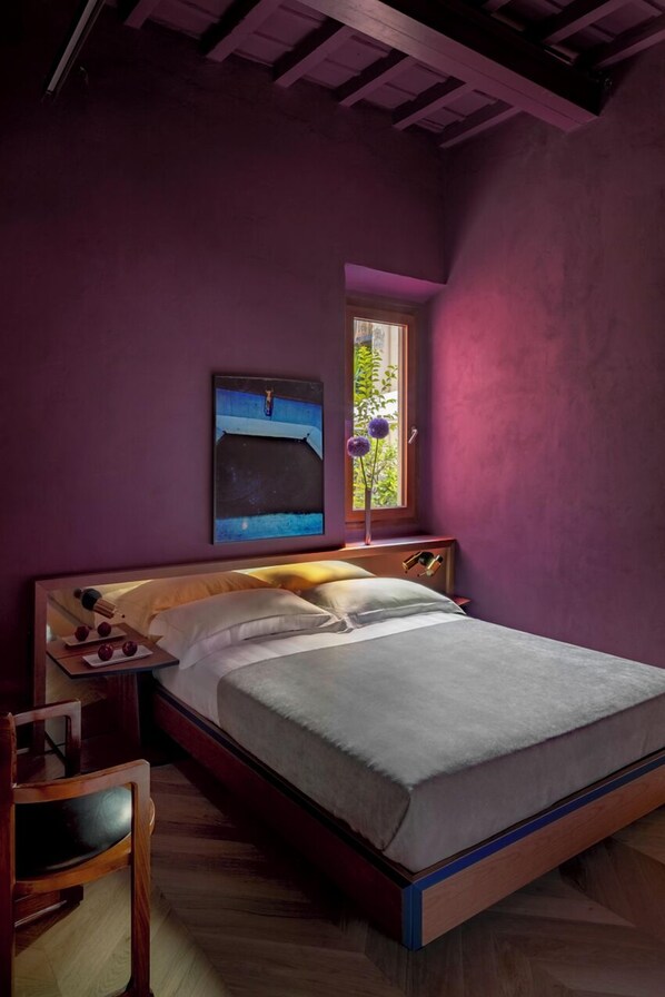 A bedroom with a bed, a nightstand, a chair, a window with a view of greenery, and a painting on the wall.