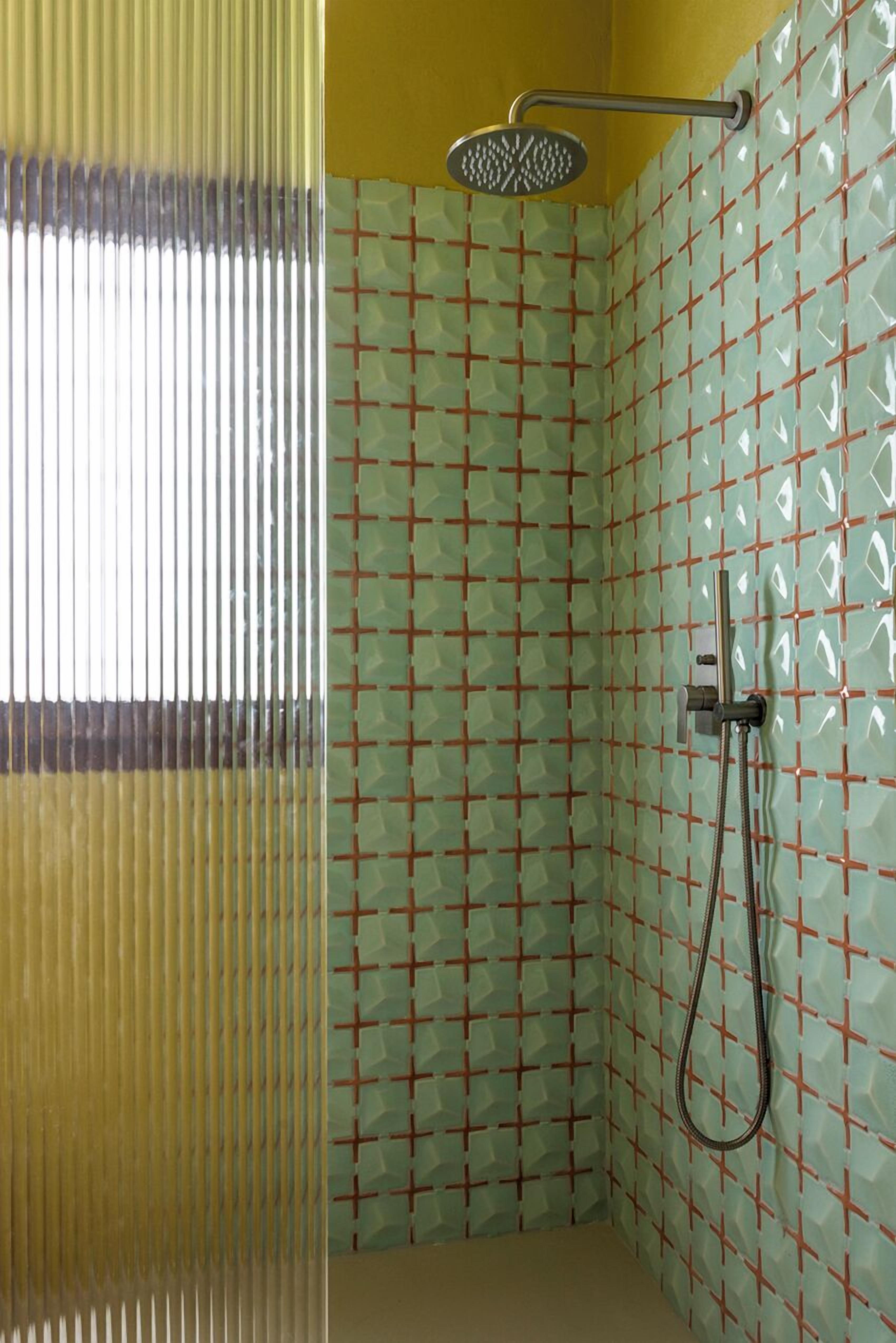 A shower with a handheld showerhead and a handheld hose, tiled walls with a geometric pattern, and a window with vertical blinds.