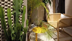 Un área de estar moderna al aire libre con una silla de mimbre y una mesa amarilla, rodeada de plantas diversas, incluyendo un cactus grande y palmeras.