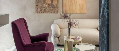 A modern living room with a purple armchair, a beige sofa, and a green patterned rug. There are abstract wooden wall art pieces and a floor lamp.