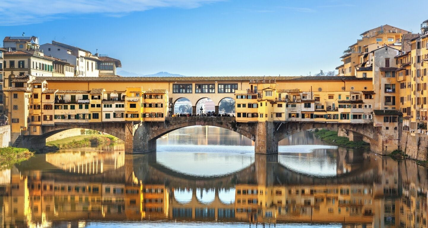 A historic bridge with multiple arches spanning a calm river, surrounded by traditional buildings with balconies and large windows.