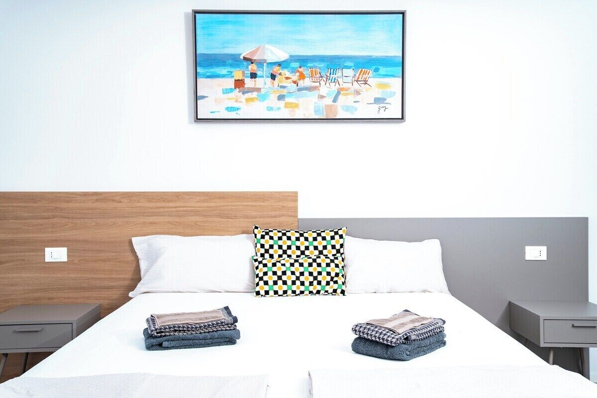 A hotel room with a bed, two pillows, a framed painting of a beach scene, and a neatly made bed with folded towels.