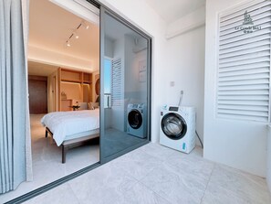 Quarto de hotel com cama, máquina de lavar e porta de correr de vidro que dá para um banheiro.