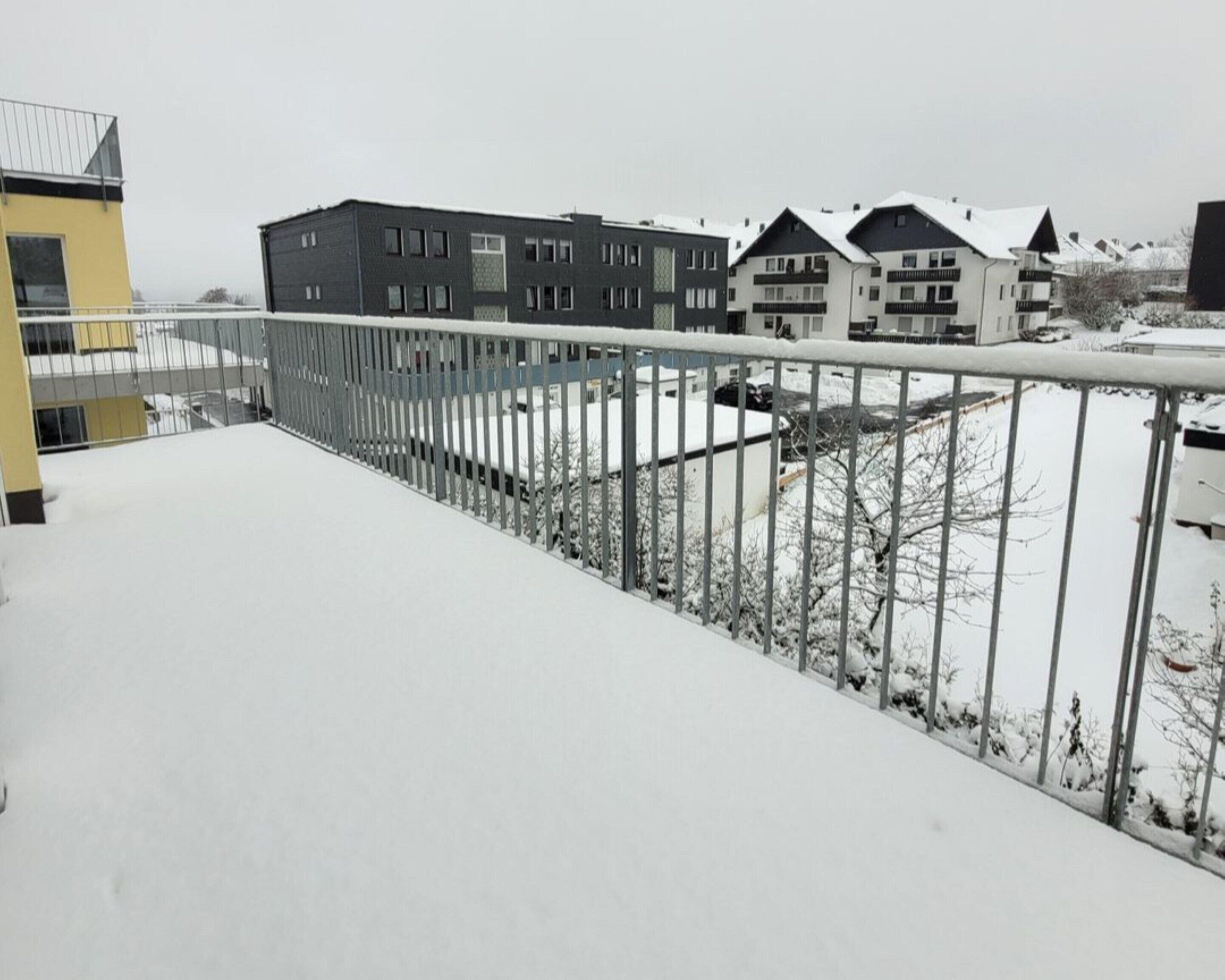 Un balcone imbiancato dalla neve con ringhiera metallica che si affaccia su una fila di edifici a più piani.