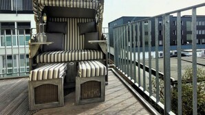 Eine Dachterrasse mit einer gestreiften Couch, zwei Beistelltischen und einem kleinen Tisch mit einem Glas.