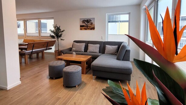 Ein modernes Wohnzimmer mit einem grauen Ecksofa, einem Holztisch und zwei grauen Hocker. Es gibt einen Essbereich mit einem Holztisch und Stühlen sowie ein großes Fenster mit Jalousien.